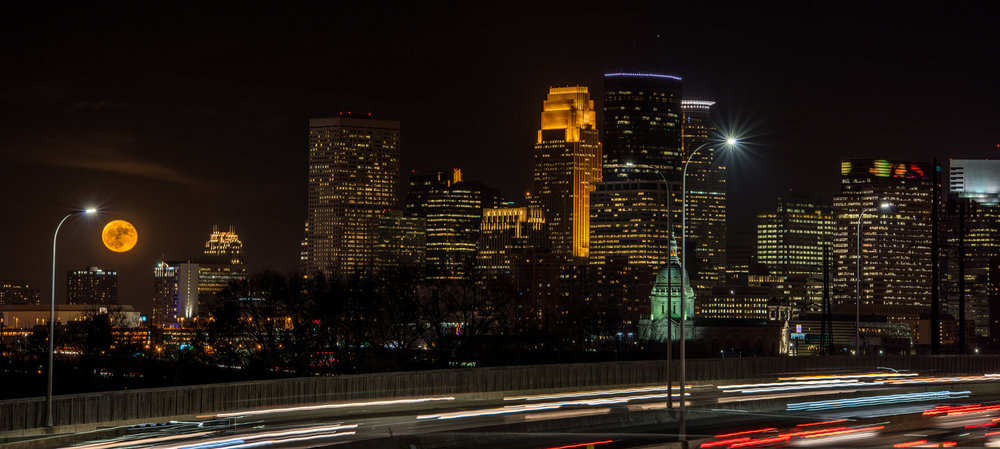 Full moon rising over a city skyline at night with blurred car lights on a highway in the foreground.