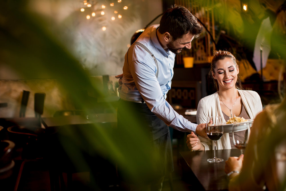 A waiter serves food to a smiling woman at a restaurant table with wine glasses.