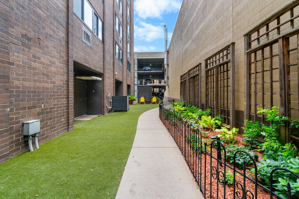 Narrow path between buildings with green grass, a small garden, and yellow chairs visible in the distance.