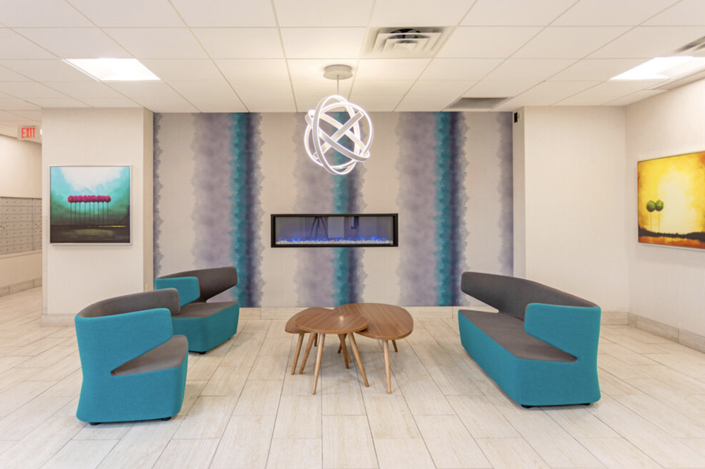 Modern lobby with teal and gray seating, a central wooden table, wall art, and an electric fireplace.