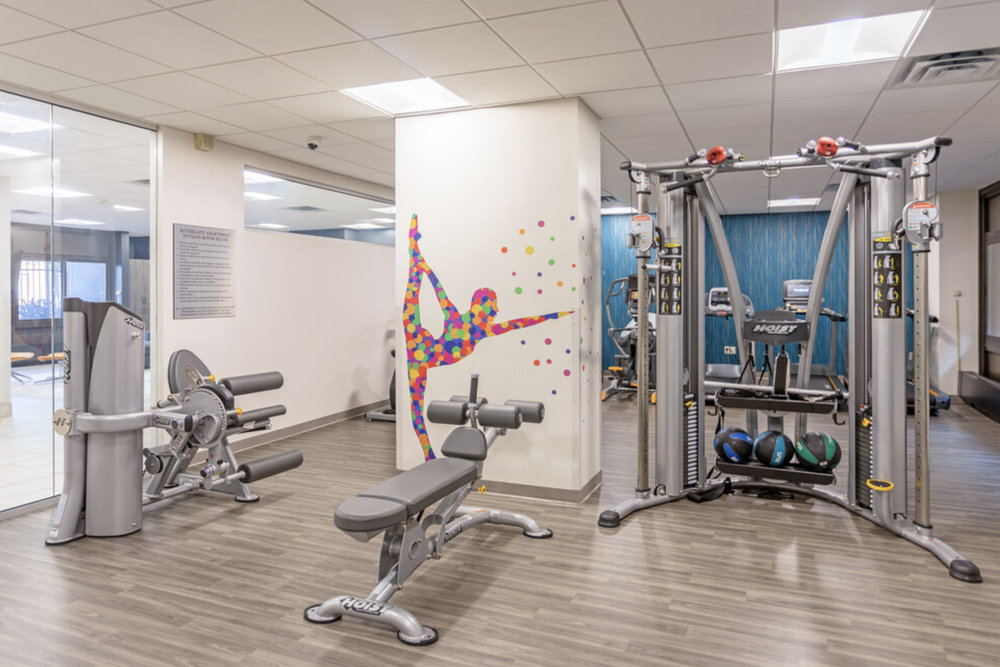 A modern gym with weight machines, a bench, medicine balls, and a colorful dancer mural on the wall.