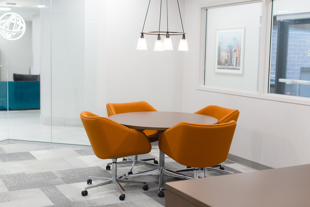 Four orange chairs surround a round table under modern lights in a bright, modern office space.