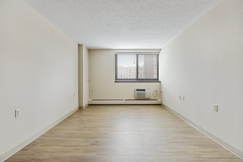 Empty room with light wood flooring, beige walls, and a window with blinds and an air conditioning unit below.