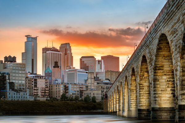 Stone arch bridge over river with city skyline and sunset in the background.