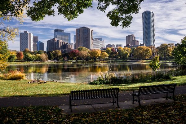 Two empty benches face a city skyline across a lake in a park, with autumn trees and scattered fallen leaves.
