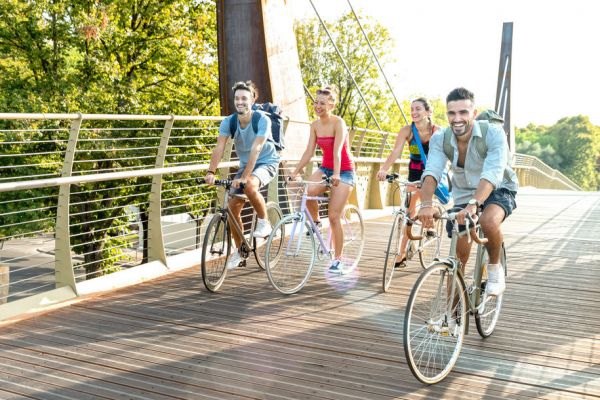 Four young adults ride bicycles together on a wooden bridge surrounded by trees, smiling and enjoying the day.