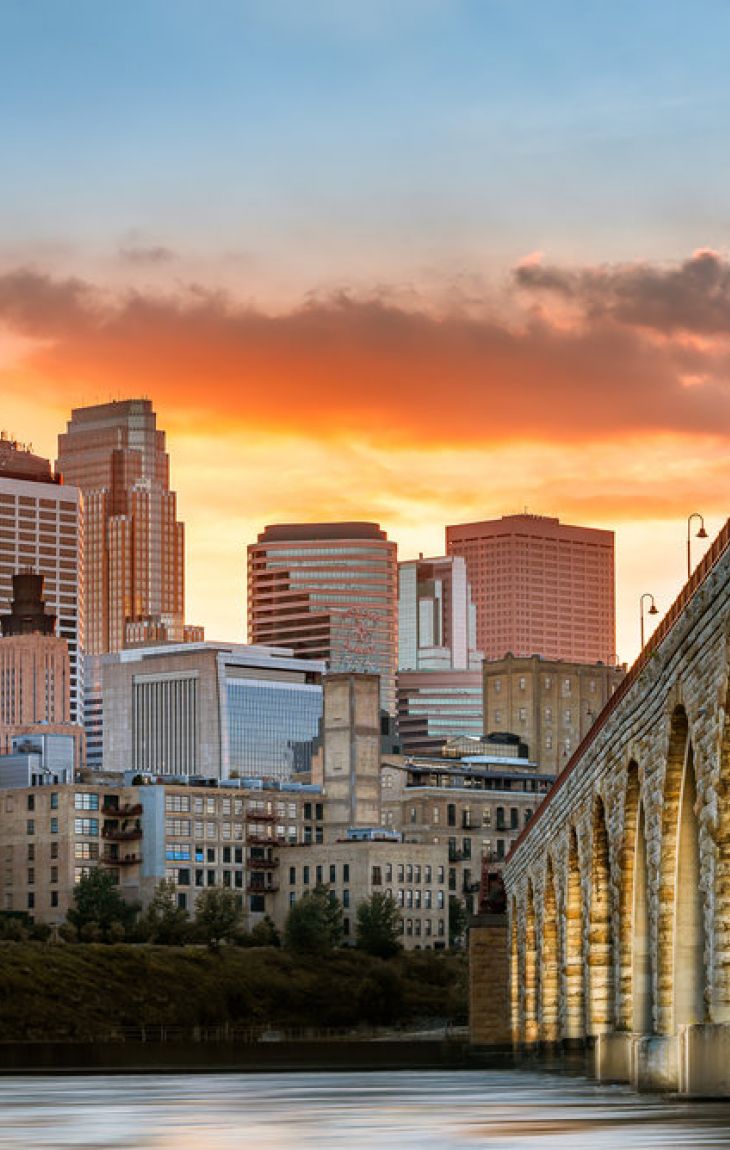 Stone arch bridge over river with city skyline and sunset in the background.