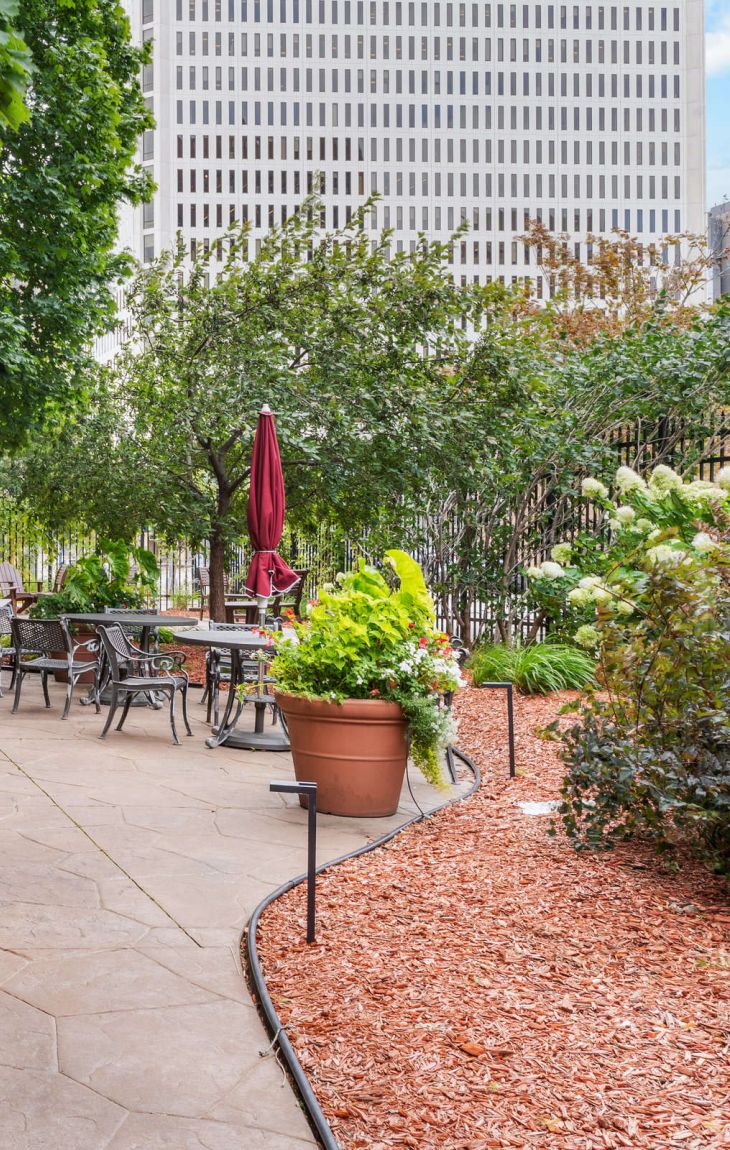 Outdoor patio with tables, chairs, grill, potted plants, and trees along a paved walkway near a city building.